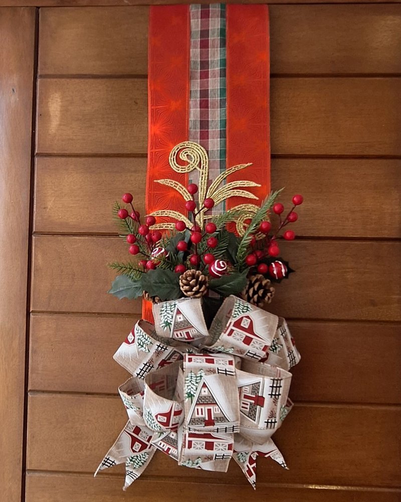 Escapulario navideño colgado en una puerta de madera: cinta roja con tartán, ramas de pino, berries, piñas y moño estampado